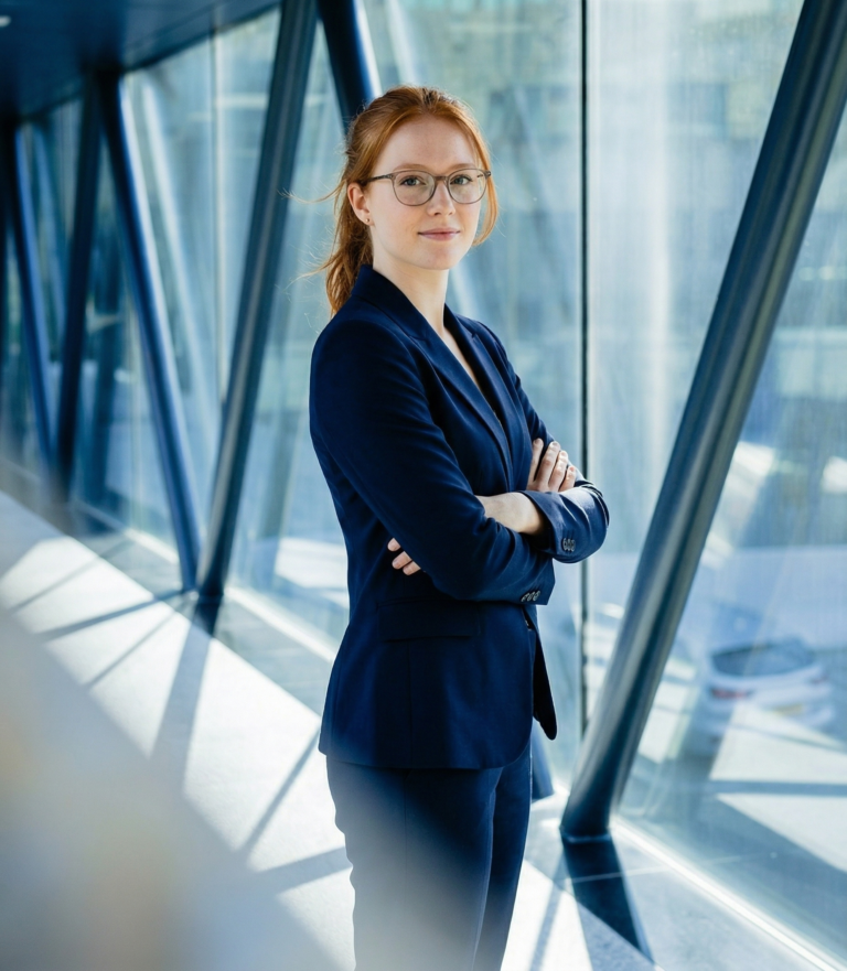 Een vrouw met een bril en gekleed in een donker blauw pak staat in een gang met veel raam. Ze staat trots met haar armen over elkaar heen en kijkt de camera in